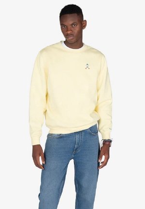 Sudadera amarilla de manga larga y cuello redondo, con un logo bordado. Combinada con jeans de mezclilla azules. Diseño sencillo.