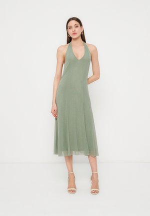 Nő áll, olívazöld halter nyakú midi ruhát és bézs nyitott orrú bokapántos magassarkút visel egyszerű fehér háttér előtt.