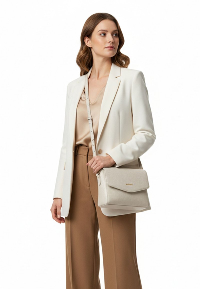 Femme portant un blazer blanc et un pantalon beige tenant un sac à bandoulière beige avec une fermeture à rabat sur fond blanc.