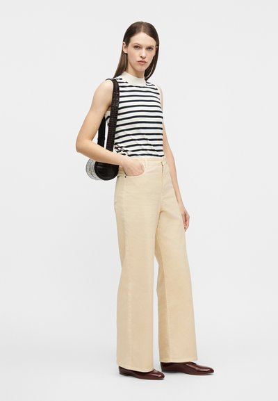 Γυναίκα με αμάνικη μαύρη και λευκή ριγέ μπλούζα, μπεζ παντελόνι φαρδιάς γραμμής, καφέ παπούτσια και μαύρη τσάντα ώμου.