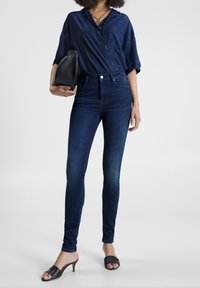 Jeans skinny de tiro alto en azul oscuro combinados con una camisa de denim y mule de tacón negro. La modelo sostiene un bolso negro. Diseño simple y ajustado.