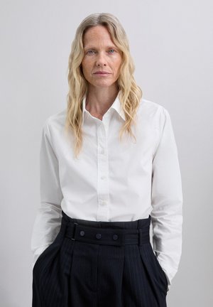 Hvid skjorte med lange ærmer og spids krave, parret med højtaljede, stribede bukser med en bred talje med knapper.