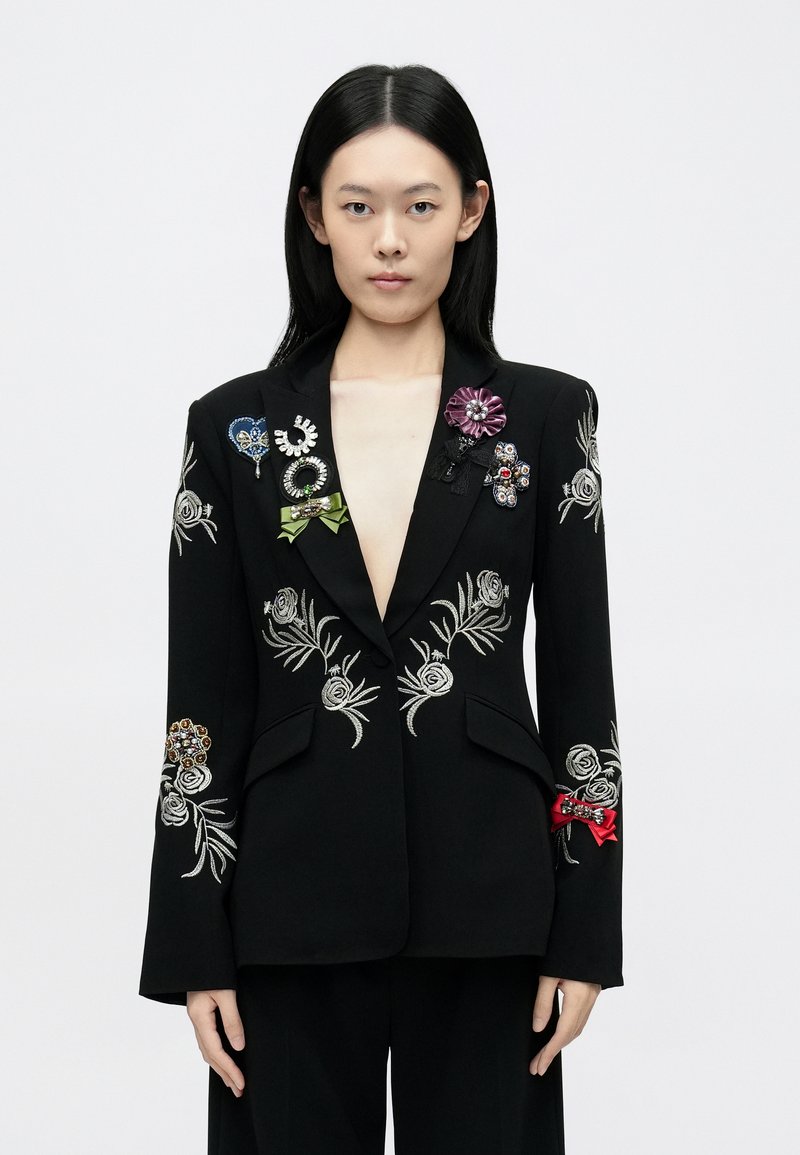 Fekete blézer ezüst virágos hímzéssel és különböző színes brossokkal díszítve. Kiemelkedő részletek a mély V-nyak és a strukturált zsebek.