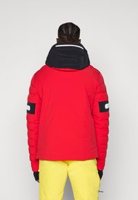 Rote, gepolsterte Jacke mit schwarzer Kapuze, mit gesteppter Textur, einem Logo auf dem Ärmel und einem schlanken Design. Gelbe Hose ergänzt das Outfit.