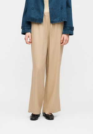 Donna che indossa pantaloni beige a gamba larga con vita elastica, giacca di jeans blu scuro e ballerine nere, in piedi davanti a uno sfondo bianco.