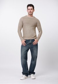 Pull beige avec un motif texturé et un col rond, associé à un jean bleu ample et des baskets blanches, sur un fond blanc uni.