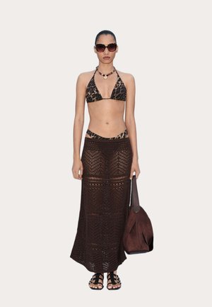 Kvinde iført leopardprint bikini top og bund, lang brun hæklet nederdel, sorte sandaler, solbriller, halskæde og holder en brun håndtaske.