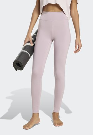 Άτομο που φοράει ανοιχτό ροζ κολάν και λευκό κοντό τοπ, κρατώντας ένα μαύρο τυλιγμένο στρωματάκι γιόγκα με το ένα χέρι, ξυπόλητο σε λευκή επιφάνεια.