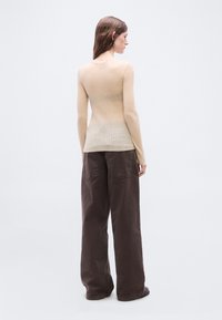 Femme aux longs cheveux bruns portant un haut beige à manches longues côtelé et un pantalon ample marron foncé, debout de dos sur un fond blanc.