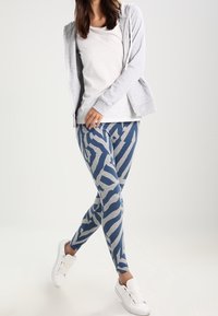Leggings a strisce zebra blu e grigie realizzati in tessuto elasticizzato, abbinati a una maglietta bianca e a una felpa grigia con zip, completati da sneakers bianche.