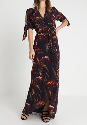 Femme portant une robe portefeuille noire à imprimé floral, manches courtes nouées au niveau des coudes, et des sandales noires à fines lanières.