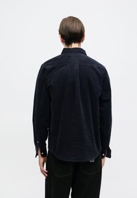 Ναυτικά μπλε πουκάμισο από κοτλέ με γιακά, κουμπιά στους καρπούς και κυρτό στρίφωμα. Διαθέτει κάθετη υφή ρίβ και λεπτομέρεια στον πίσω γιακά.