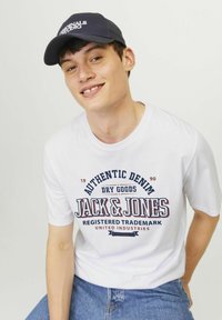 Hvid t-shirt med korte ærmer, der har rød og marineblå teksttryk "Authentic Denim" og "Jack & Jones." Mørkeblå kasket med hvidt logo. Denimshorts.