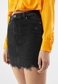 Minigonna in denim nero con orlo sfrangiato, design a cinque tasche e chiusura frontale con bottoni. Abbinata a una camicia arancione brillante.