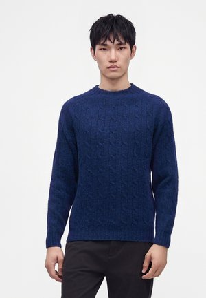 Homme portant un pull en maille torsadée bleu foncé et un pantalon noir, debout devant un fond clair uni.