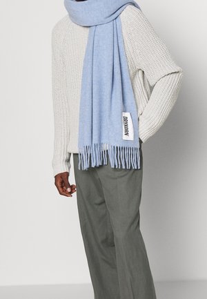 Personne portant une écharpe bleu clair avec une étiquette "DRYKORN" sur un pull en maille crème et un pantalon gris, debout avec une main sur la hanche.