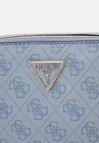 Modrá tkanina s logem Guess, zdobená vyraženým vzorem, se stříbrným trojúhelníkovým štítkem, zapínáním na zip a strukturovaným povrchem.