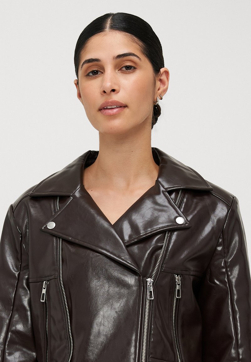 Veste de moto en cuir marron avec grands revers, fermetures éclair argentées et deux poches zippées, présentant une finition brillante et un design structuré.