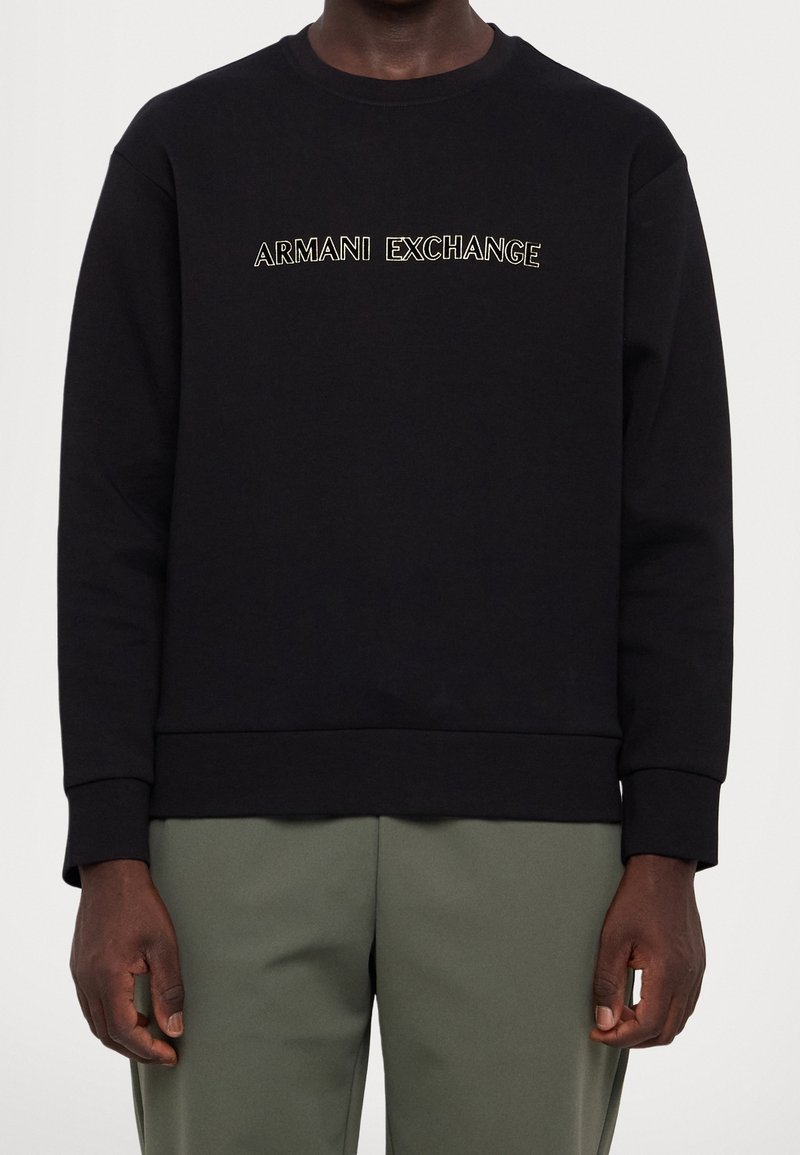 Homme portant un sweat-shirt noir avec le texte "ARMANI EXCHANGE" en blanc sur la poitrine et un pantalon vert olive.