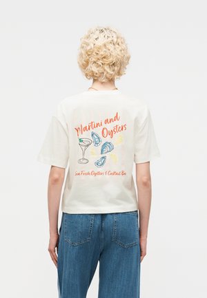 Personne aux cheveux blonds courts et bouclés de dos, portant un T-shirt blanc avec le texte « Martini and Oysters » et des illustrations de martini, huîtres et quartiers de citron.