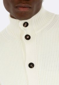Maglione bianco a coste con colletto alto, caratterizzato da tre bottoni scuri e un motivo testurizzato sul tessuto. Vista ravvicinata della sezione superiore.