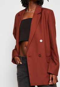Personne portant un blazer couleur rouille sur un top bandeau noir et un jean gris foncé, avec une main dans la poche et un collier chaîne argenté.