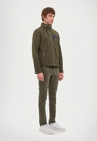 Mees seisab, seljas oliiviroheline kapuutsiga jakk, sellele sobivad püksid ja valged tossud, seistes vasakule, eine lihtsa heleda taustaga.