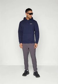 Un homme portant un hoodie bleu marine, un pantalon de jogging gris, des baskets noires et des lunettes de soleil noires se tient devant un fond blanc uni.