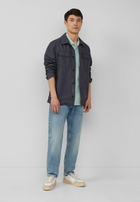 Donker marineblauwe button-up jas met twee borstzakken, lichte mintgroene shirt eronder, lichtblauwe jeans en witte sneakers met beige accenten.