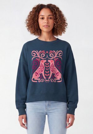 Frau mit lockigem Haar trägt einen dunkelblauen Pullover mit symmetrischem pinkfarbenem Hundedesign mit Blumen- und Wirbelmustern, kombiniert mit hellen Jeans.