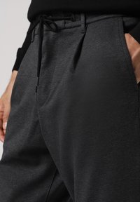 Černé kalhoty s vzorem a texturovanou tkaninou, s elastickým pasem na šňůrku a bočními kapsami. Design zahrnuje přizpůsobený střih.