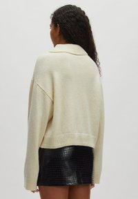 Gekürzter, cremefarbener Pullover aus weichem Strick mit Schalkragen, kombiniert mit einem glänzenden, schwarzen Leder-Minirock mit Alligator-Textur.