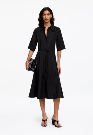 Femme portant une robe midi noire boutonnée avec des manches mi-longues, tenant une pochette noire et portant des sandales à talons noires.