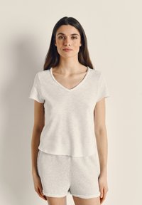 Sieviete ar gariem tumšiem matiem, valkājot baltu īsroku V veida kakla topu un gaiši pelēkas šortus, stāv pret vienkāršu gaišu fonu.