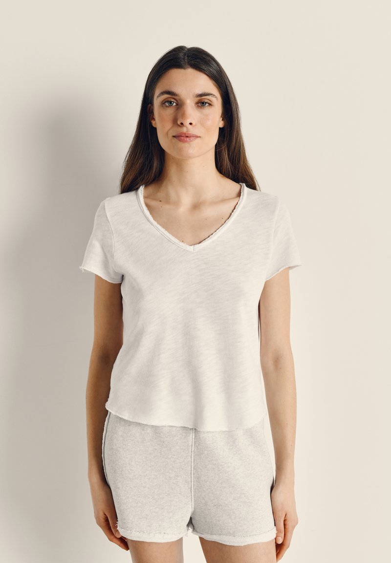 Sieviete ar gariem tumšiem matiem, valkājot baltu īsroku V veida kakla topu un gaiši pelēkas šortus, stāv pret vienkāršu gaišu fonu.