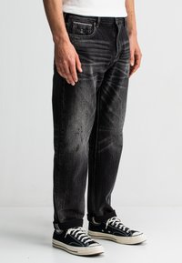 Schwarze Jeans aus Denim mit einem verblassten Finish, geradem Schnitt und subtilen, gewollten Verwaschungen. Kombiniert mit schwarzen Segeltuchschuhen mit weißen Akzenten.