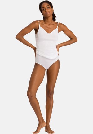 Femme debout pieds nus, mains sur les hanches, portant un caraco blanc et une lingerie assortie sur un fond uni.