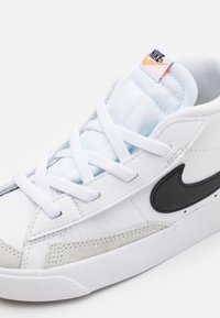 Bílé tenisky s texturovaným koženým svrškem, šedými semišovými doplňky a černým logem Nike. Mají světle modrou podšívku a bílé tkaničky.