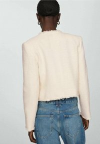 Cremefarvet cropped jakke lavet af textureret stof med afrivende kanter. Sat sammen med blå højtaljede jeans, som viser bagsiden.