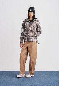 Pulloverjack met een gemêleerd bruin en grijs patroon, zwarte muts, beige broek met wijde pijpen en grijze sneakers met lichte accenten.