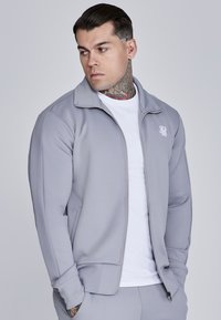 Giacca zip grigia chiara con colletto alto e logo, abbinata a una maglietta bianca sotto. Tessuto morbido con design aderente.