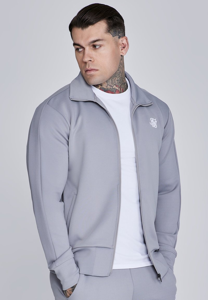 Giacca zip grigia chiara con colletto alto e logo, abbinata a una maglietta bianca sotto. Tessuto morbido con design aderente.
