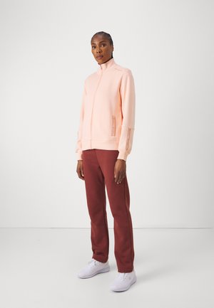 Giacca in felpa rosa chiaro con zip e colletto alto, dotata di tasche laterali e polsini a costine, abbinata a pantaloni della tuta bordeaux e sneakers bianche.