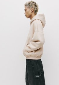 Felpa beige con tasca frontale e cappuccio con lacci, abbinata a pantaloni cargo grigi scuri con una tasca laterale etichettata "STRUMENTO".