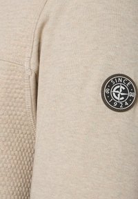 Beige sweatshirt met een textuur, voorzien van een ronde zwarte patch met het opschrift "SINCE 1924" op de linkermouw.