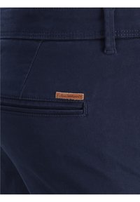 Pantalones azul oscuro con un bolsillo trasero de ojal que presenta una pequeña etiqueta de cuero marrón cosida encima de la abertura del bolsillo.