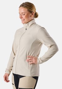 Veste en polaire beige avec un col haut, fermeture éclair avant intégrale et deux poches latérales. Texture lisse et design ajusté avec un détail de marque discret.