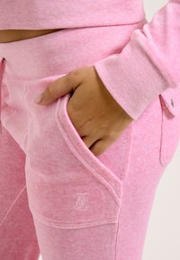 Rosa Fleece-Jogginghosen mit strukturiertem Finish, ausgestattet mit schrägen Taschen und einem kleinen Logo an der Seite. Weicher Stoff und lockere Passform.
