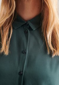 Donkergroene overhemd met kraag, gemaakt van een gladde stof. Heeft zwarte knopen in het midden en een blousachtige textuur.