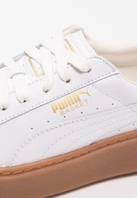 Baskets Puma blanches avec logo doré, lacets blancs, couture détaillée et semelle en caoutchouc marron, montrées en gros plan.